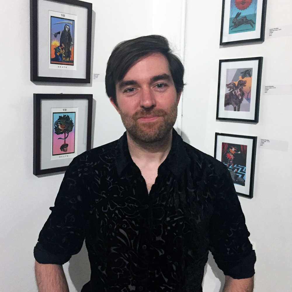 Photograph of Andrew Wheatley stood in front of a gallery wall of his framed illustrations.